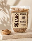 A clear stand-up pouch of Gran Mitla Sal de Gusano (Gusano Warm Salt with chili) displayed on a rustic wooden table alongside a small bowl of seasoning, a glass of clear liquid, and a dark ceramic vase with agave leaves in the background.