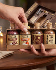 A gift set of four small glass bottles with cork stoppers, labeled Jamaica, Habanero, Chapulín, and Graso, presented in a kraft paper box held by two hands over a wooden table. An open matching box with a glass and spoon is visible behind it, along with a wine glass on the table.