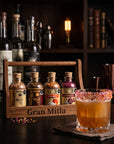 Four small artisanal spice bottles in a wooden carrier labeled 'Gran Mitla' on a dark bar counter, with a garnished amber drink in a rocks glass in the foreground and bottles and shelves blurred in the background.