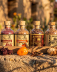 Four glass spice bottles with cork stoppers from Gran Mitla are arranged on a stone surface outdoors. The bottles contain colorful dried spices and are labeled Jamaica, Habanero, Chapulín, and Susano.
