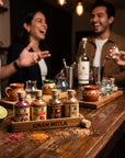 A group of people laughing around a rustic wooden bar counter, with several small bottles of Gran Mitla spices and herbs arranged in a branded wooden tray. The scene is warmly lit by hanging Edison bulbs, with limes, mixing tools, and other ingredients visible on the counter.