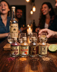 A group of people gathered around a wooden table in a warmly lit bar or kitchen setting, smiling and laughing while drinking from small glass bottles of spices and herbs. In the foreground, four labeled bottles with cork stoppers sit on the table, with loose spice blends scattered in front of them and a lime half visible on the right.