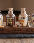 Four glass spice bottles with cork stoppers and printed labels are arranged on a wooden tray engraved with spice names, alongside a small spoon, on a dark stone serving board over a wooden table.