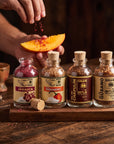 Four glass bottles of Gran Rio Grano spices on a wooden tray, with a person's hand sprinkling red chili powder onto a slice of mango. The bottles are labeled Jamaica, Habanero, Jal de Chaplin de Nitla, and El Guayabo.