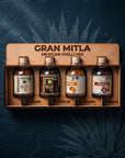 A boxed set of four Gran Mitla Mexican Chilli Mix bottles displayed in a wooden tray against a dark textured background with leaf silhouettes. The bottles are labeled Gusano, Chapulin, Habanero, and Jamaica.