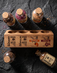 A wooden spice rack holding four glass spice bottles with cork stoppers, arranged on a dark stone surface. One bottle is upright in the center, two are on the left, and one lies on its side at the bottom right.