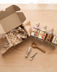 An open kraft cardboard gift box filled with brown paper shred sits on a light wood table beside four small glass bottles with cork stoppers and cream labels, along with a wooden spoon, a rolled paper item tied with string, and a small metal tool.