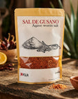 A stand-up pouch of Mexa Sal de Gusano (Agave worm salt) displayed on a rustic wooden table with chili peppers, a lemon slice, and green plants in the background.