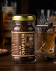 A glass jar of Sal de Chapulín (pepper) with a gold lid and a brown label, set on a wooden bar table with whiskey glasses and bottles in the background.