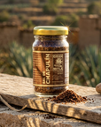A glass jar of Sal de Chapulín Gran Milla spice blend with a gold lid and brown label, set on a rustic wooden board outdoors with agave plants and clay structures in the background.