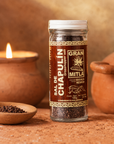 A glass jar of Gran Mitla Sal de Chapulín (grasshopper salt) set on a rustic terracotta surface with a lit candle and clay pot in the background.