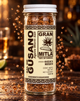 A clear glass jar of Gran Mitla Agave Worm Salt seasoning, featuring a cream-colored label with black text and a white screw cap, set against a dark, warm, blurred background with floating seasoning crystals.