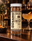 A bottle of Gran Mitla Agave Worm Salt seasoning stands on a dark wooden bar counter, with a blurred cocktail and bar shelves in the background.