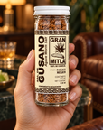 A hand holding a clear glass jar of Gran Mitla Agave Worm Salt seasoning, showing the textured brown seasoning inside, with a blurred warm-toned indoor background.