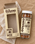 A glass jar of Gran Mitla Sal de Gusano agave worm salt next to its kraft paper packaging box, both featuring a vintage-style cream and brown label, set on a brown paper background with white tissue paper.