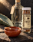 A jar of Gusano Agave Worm Salt seasoning next to a glass bottle and a small terracotta bowl with a spoon, set on a rustic stone surface with natural sunlight and shadows.