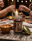 A jar of Gran Milto Sal de Chapulin coffee sits on a rustic wooden table at a dinner table, with people holding glasses in the background. A small clay bowl with a yellow drink and a gold lid are also on the table.