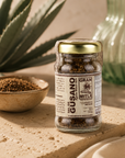 A glass jar of Gran Mitla Gusano Alawa Mole Salt with a gold lid and brown label, set on a stone surface next to a small bowl of the ground salt, with an aloe vera plant and a green glass bottle in the background.