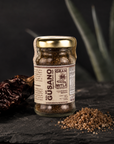 A glass jar of Gran Mitla Guasano seasoning blend with a gold lid, set on a dark slate with dried chiles and seasoning powder. The background features blurred green aloe leaves.