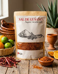 A stand-up pouch of Sal de Gusano (Agave worm salt) by the brand Mexa, styled on a rustic wooden table with ingredients like dried red chilies, limes, oranges, and a terracotta cup.