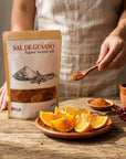 A person in a beige linen apron stands behind a wooden table, holding a wooden spoon with orange-red seasoning above a plate of sliced oranges. To the left is a stand-up pouch labeled 'SAL DE GUSANO Agave worm salt' with the brand 'Meya' visible near the bottom.