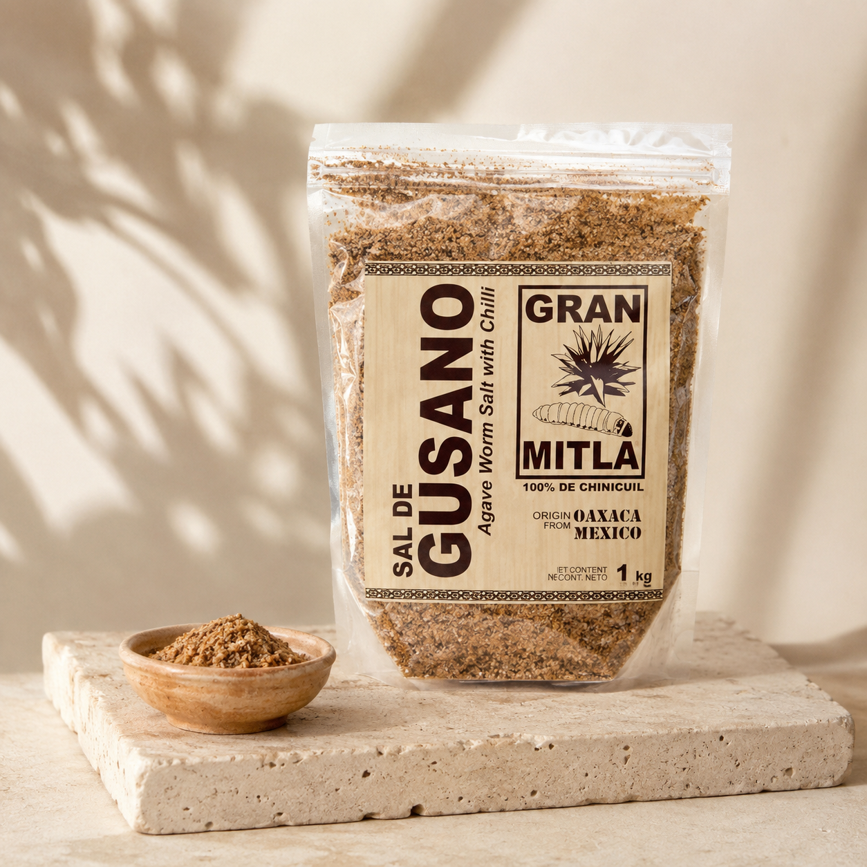 
A clear stand-up pouch of Gran Mitla Sal de Gusano (Gusano Warm Salt with chili) displayed on a rustic wooden table alongside a small bowl of seasoning, a glass of clear liquid, and a dark ceramic vase with agave leaves in the background.