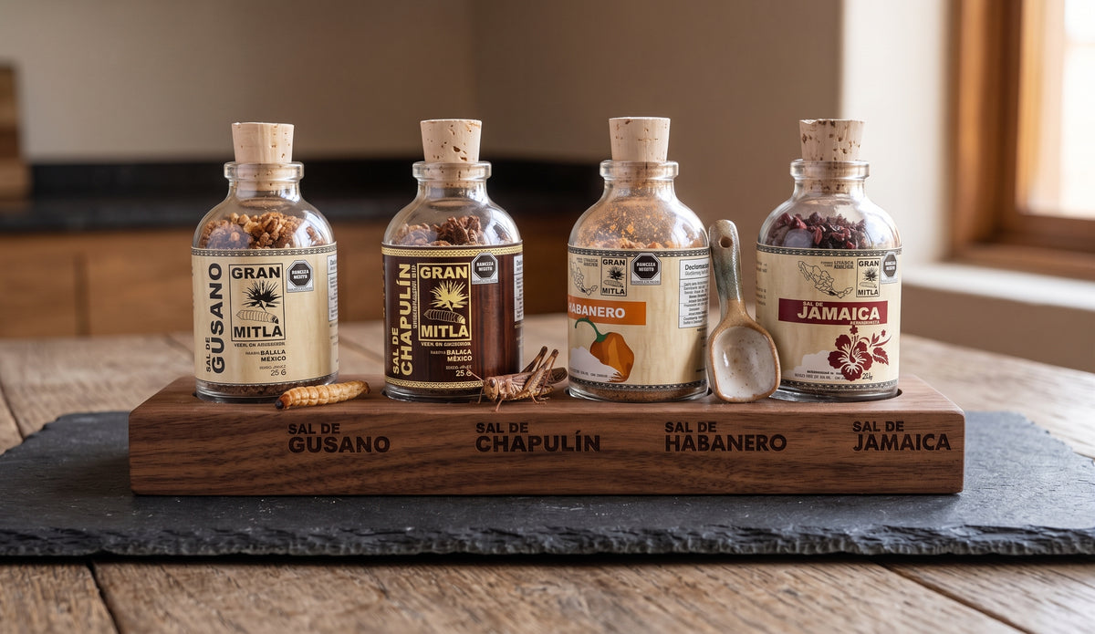 Four glass spice bottles with cork stoppers and printed labels are arranged on a wooden tray engraved with spice names, alongside a small spoon, on a dark stone serving board over a wooden table.