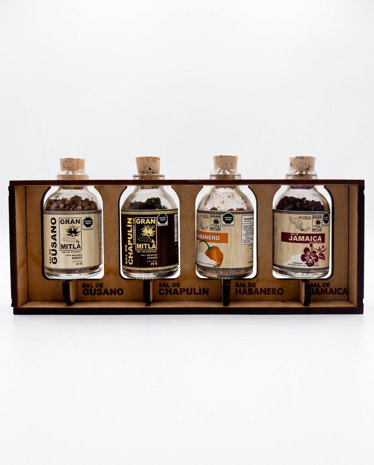 Four small glass bottles of spices displayed in a wooden tray. The bottles are labeled Gran Milla, Chaplin Milla, Habanero, and Jamaica, with cork stoppers and clear glass bodies showing the spices inside.