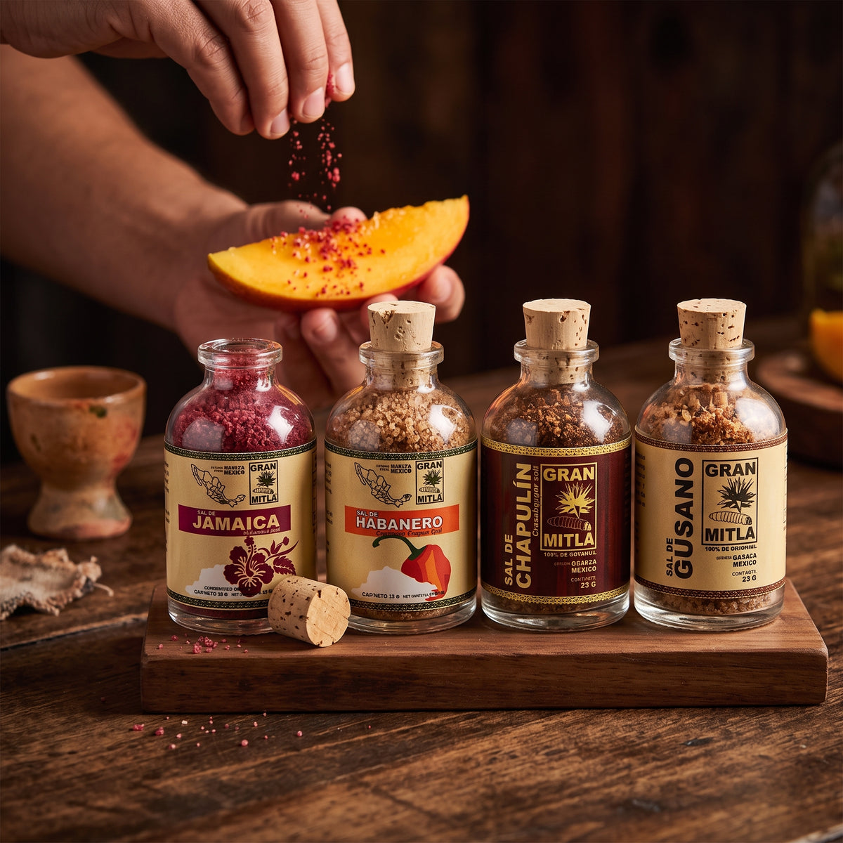 Four glass bottles of Gran Rio Grano spices on a wooden tray, with a person's hand sprinkling red chili powder onto a slice of mango. The bottles are labeled Jamaica, Habanero, Jal de Chaplin de Nitla, and El Guayabo.