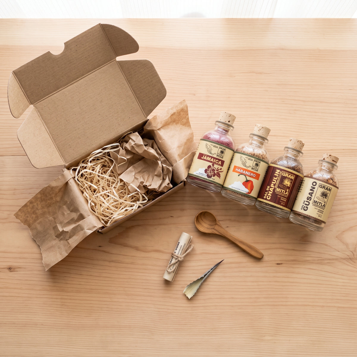 An open kraft cardboard gift box filled with brown paper shred sits on a light wood table beside four small glass bottles with cork stoppers and cream labels, along with a wooden spoon, a rolled paper item tied with string, and a small metal tool.