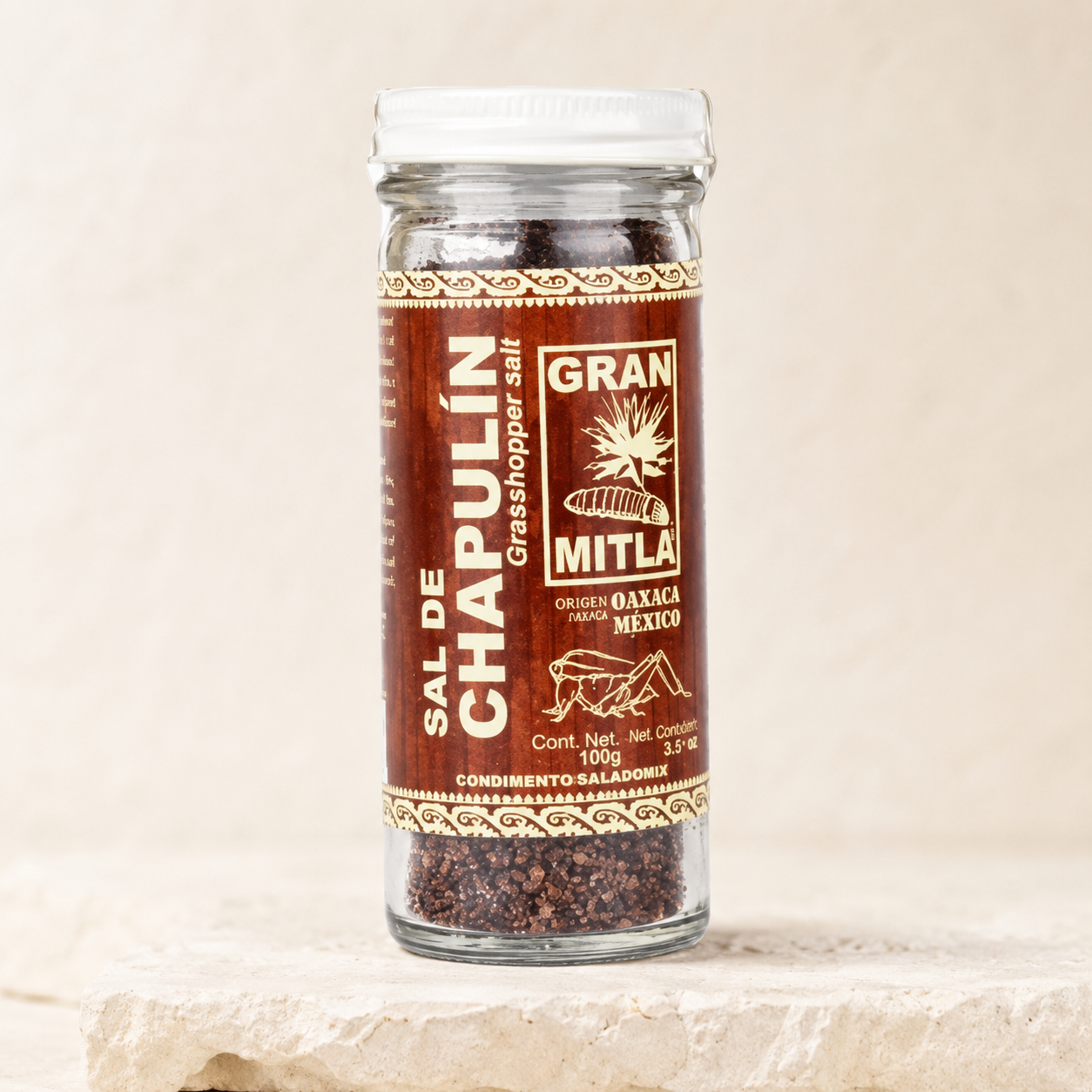 A glass jar of Mitla Sal de Chapulín grasshopper salt sits on a light beige stone surface against a matching beige background. The jar has a white lid and a brown label with gold and white text.