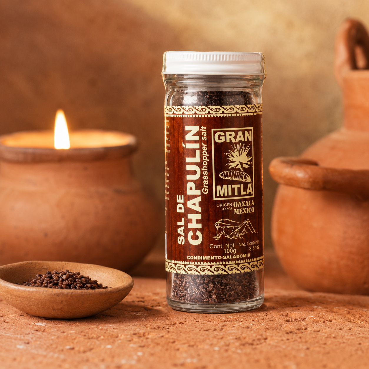 A glass jar of Gran Mitla Sal de Chapulín (grasshopper salt) set on a rustic terracotta surface with a lit candle and clay pot in the background.