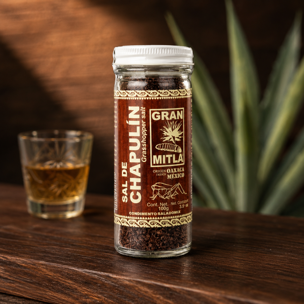 A glass jar of Sal de Chapulín Gran Mitla grasshopper salt sits on a dark wooden table next to a small glass filled with the salt, with a blurred green plant in the background.