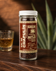 A glass jar of Sal de Chapulín Gran Mitla grasshopper salt sits on a dark wooden table next to a small glass filled with the salt, with a blurred green plant in the background.