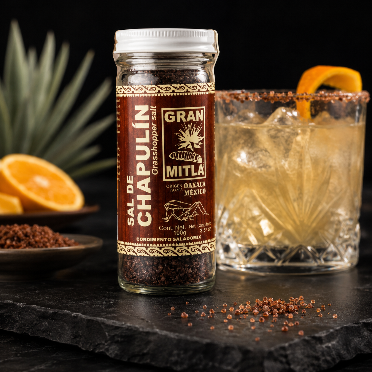 A bottle of Gran Mita Sal de Chapulin stands on a dark slate surface next to a glass of iced cocktail with a salted rim and an orange peel garnish, with fresh oranges and a pineapple in the background.
