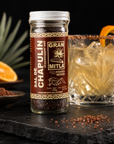 A bottle of Gran Mita Sal de Chapulin stands on a dark slate surface next to a glass of iced cocktail with a salted rim and an orange peel garnish, with fresh oranges and a pineapple in the background.