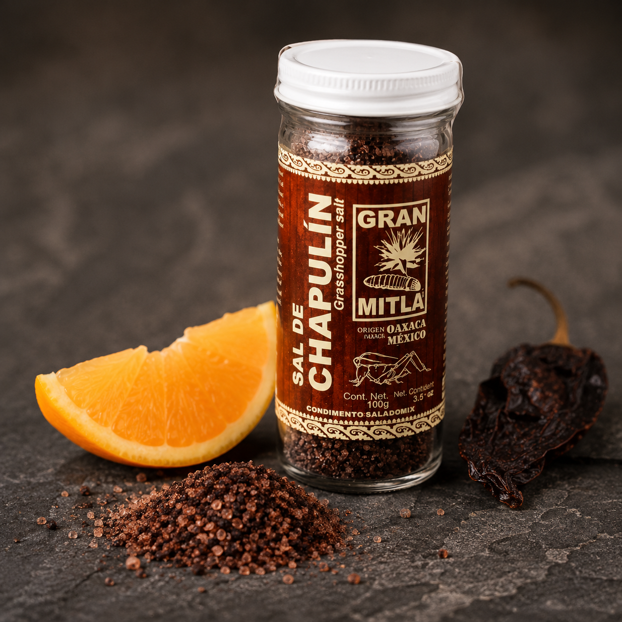 A glass jar of Chapulín grasshopper salt with a white lid and a brown label, styled on a dark textured surface with a pile of the salt, a dried chili pepper, and an orange slice.