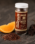 A glass jar of Chapulín grasshopper salt with a white lid and a brown label, styled on a dark textured surface with a pile of the salt, a dried chili pepper, and an orange slice.