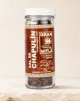 A glass jar of Mitla Sal de Chapulín grasshopper salt sits on a light beige stone surface against a matching beige background. The jar has a white lid and a brown label with gold and white text.