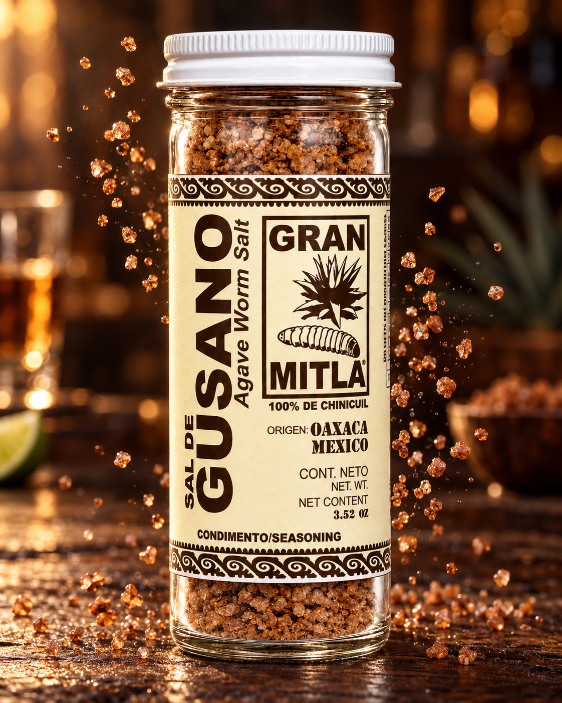 A clear glass jar of Gran Mitla Agave Worm Salt seasoning, featuring a cream-colored label with black text and a white screw cap, set against a dark, warm, blurred background with floating seasoning crystals.