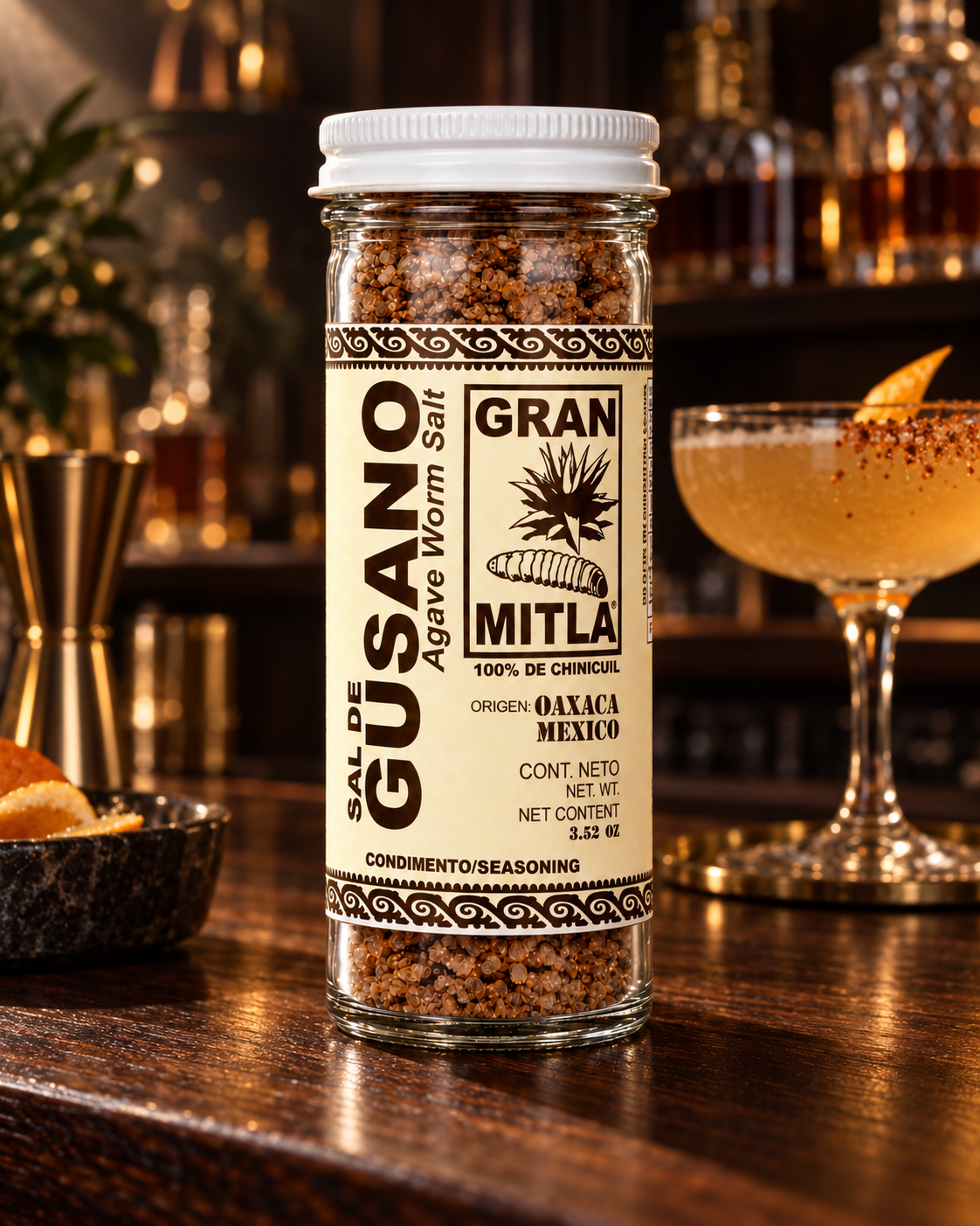 A bottle of Gran Mitla Agave Worm Salt seasoning stands on a dark wooden bar counter, with a blurred cocktail and bar shelves in the background.
