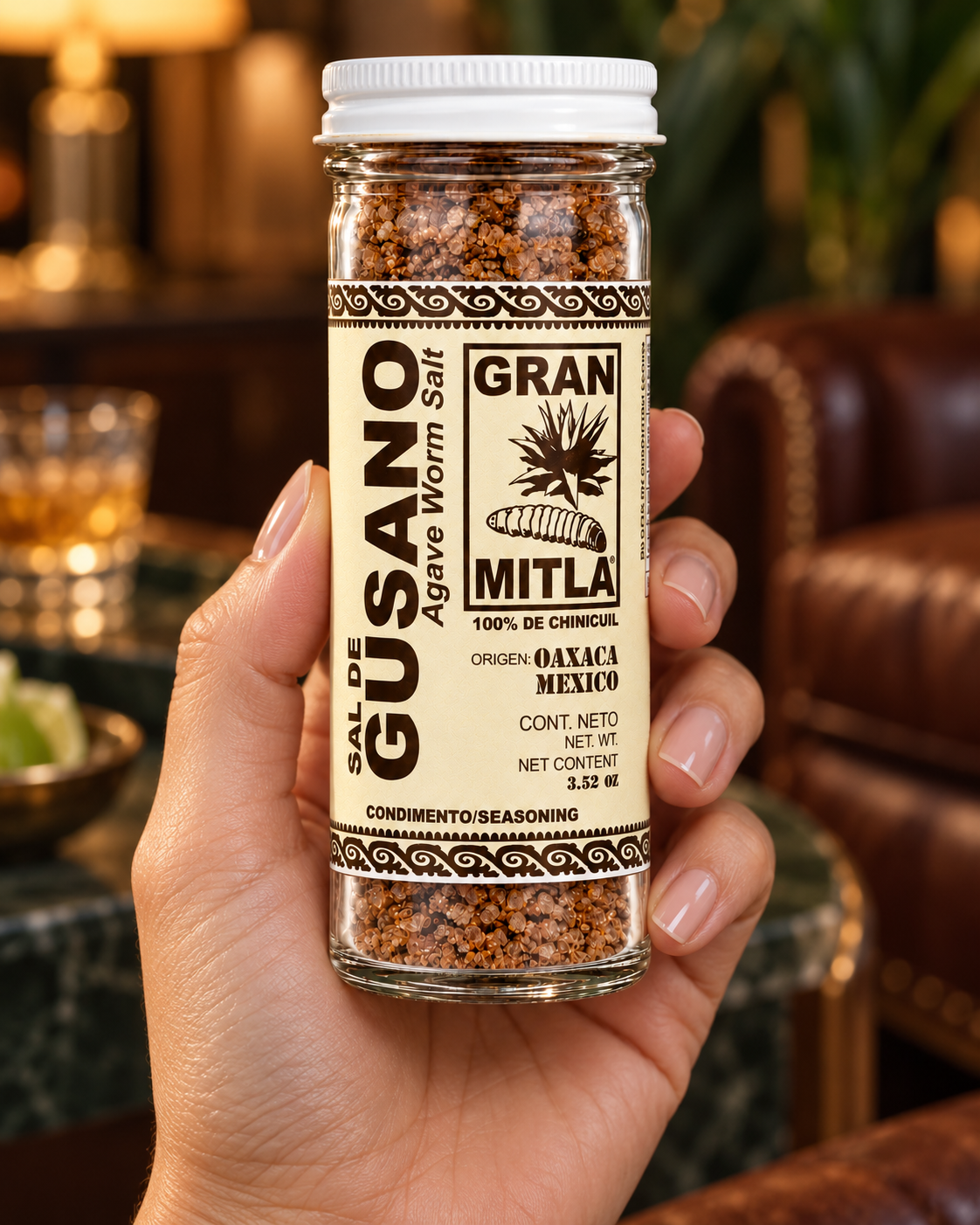 A hand holding a clear glass jar of Gran Mitla Agave Worm Salt seasoning, showing the textured brown seasoning inside, with a blurred warm-toned indoor background.