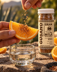 A jar of Gran Mitla Agave Worm Salt seasoning is shown next to a glass bowl of clear liquid and a hand holding a fresh orange slice, with another hand sprinkling the seasoning over the orange slice.