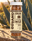 A clear glass jar of Gran Mitla Agave Worm Salt seasoning, filled with brown salt crystals, standing on a stone surface outdoors with agave plants in the background.