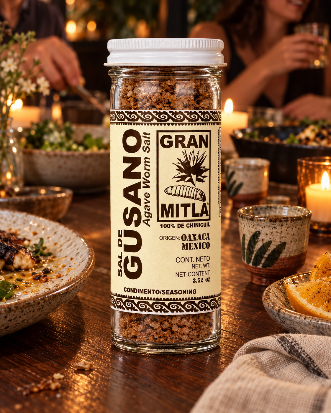 A bottle of Gran Mitla agave worm salt seasoning stands on a rustic wooden table in a warmly lit dining setting, with plates, candles, and people blurred in the background.