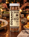 A bottle of Gran Mitla agave worm salt seasoning stands on a rustic wooden table in a warmly lit dining setting, with plates, candles, and people blurred in the background.