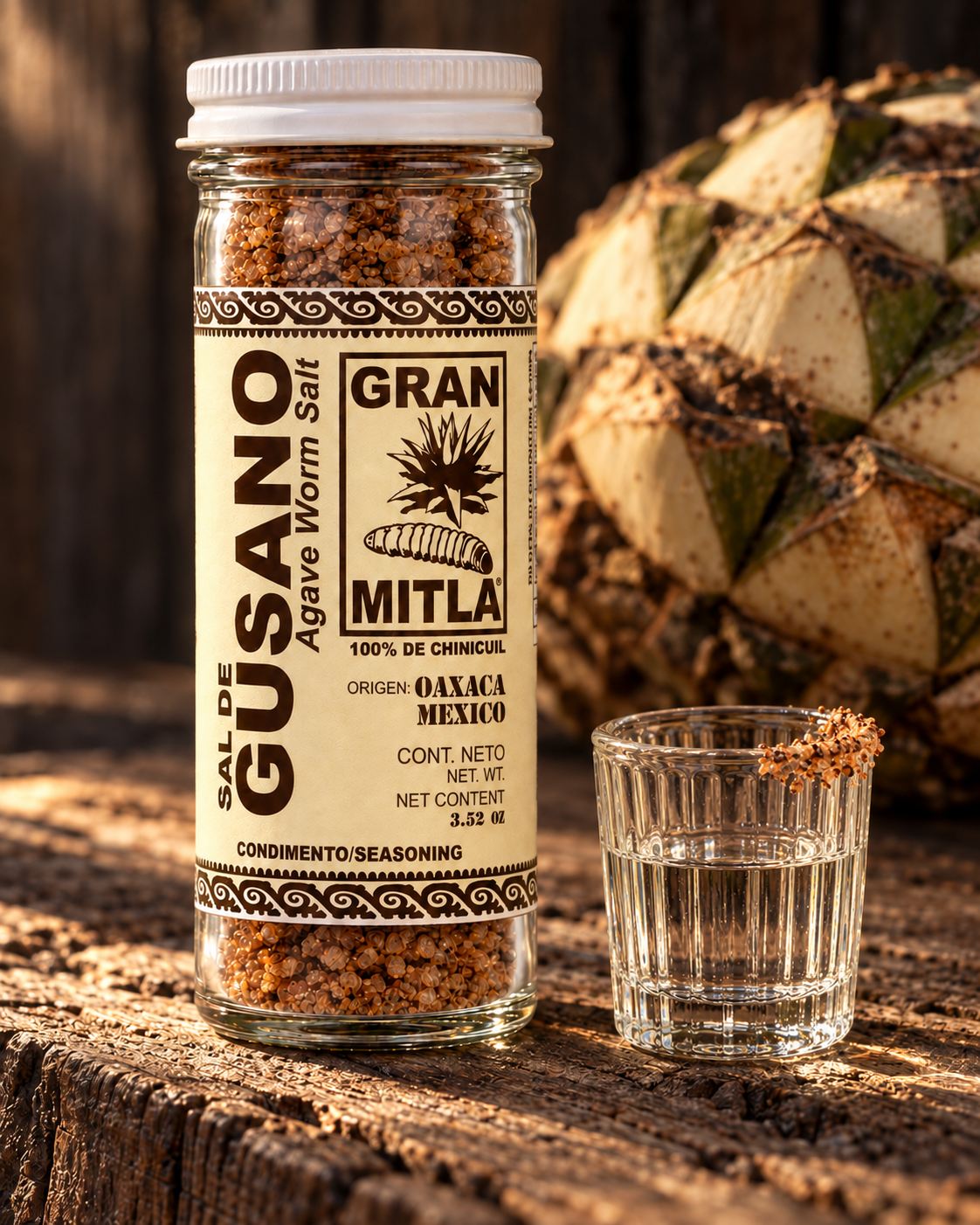 A clear glass jar of Gran Mitla Agave Worm Salt sits on a rustic wooden surface next to a small glass of the salted liquid, with a large agave cactus in the background.
