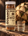 A clear glass jar of Gran Mitla Agave Worm Salt sits on a rustic wooden surface next to a small glass of the salted liquid, with a large agave cactus in the background.