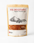 A stand-up pouch of Mexa brand Sal de Gusano (Agave Worm Salt) on a clean white background. The packaging features a kraft paper border, a white center label with red and black text, and a clear window showing the reddish-brown salt inside.