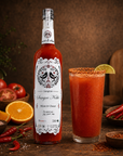 A bottle of Sangrita hot chili sauce stands next to a tall glass of the red sauce, styled on a rustic wooden table with tomatoes, oranges, chili peppers, and spices.